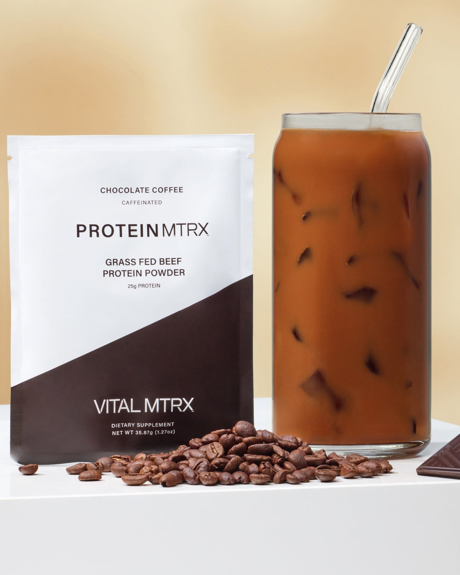 Vital MTRX protein powder package and a glass of iced protein coffee with coffee beans and chocolate pieces on a white surface.